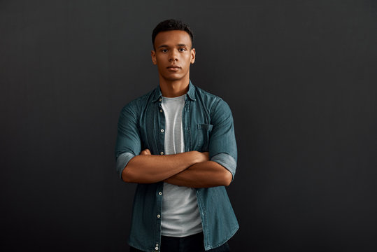 Perfect manager. Handsome and young afro american man looking at camera and keeping arms crossed while standing against dark background