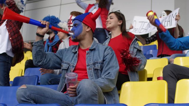 Medium shot of group of devoted young football fans sitting at stadium, watching game and supporting team with banners, pompoms and vuvuzelas, then jumping up from seats to celebrate successful goal