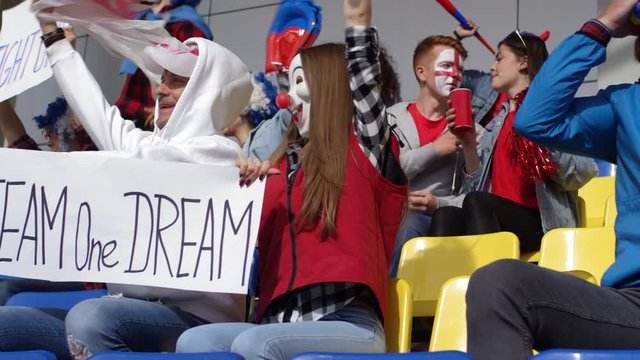 Thigh-up shot of happy young couple of fans, wearing clown masks, sitting at stadium and supporting team while holding slogan banner and waving pompons, and other excited spectators cheering around