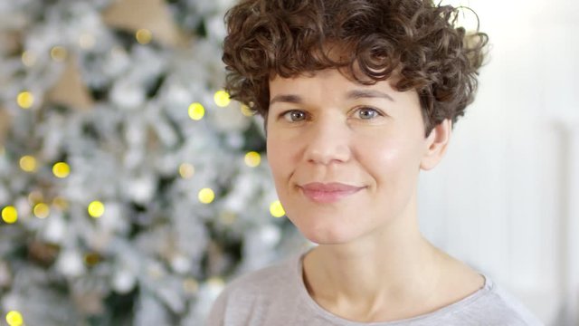 Portrait of beautiful caucasian woman with short curly hair looking at camera and smiling while posing beside Christmas tree decorated with fairy lights