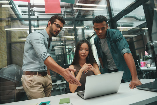 Teamwork. Young handsome bearded man pointing at computer monitor and discussing project with his two colleagues while working in modern office