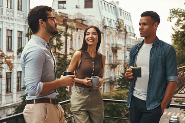 Coffee break. Young cheerful colleagues in casual wear holding cups and talking with each other while standing on the office balcony