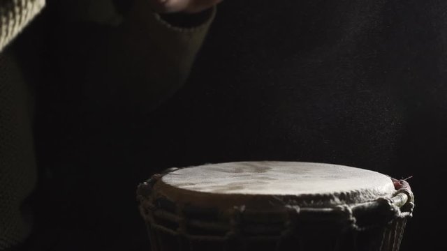 A man playing the djembe drum covered with talcum powder on the dark background, slow motion 2x, side view