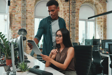 Office life. Young afro american man in casual clothes showing reports on his digital tablet and discussing new project with his beautiful female coworker