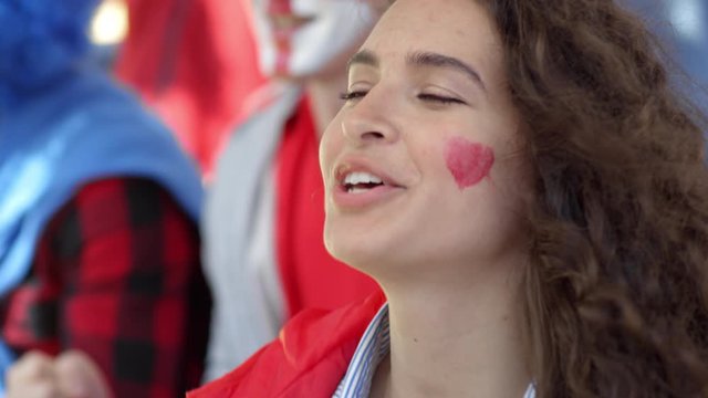 Close-up face shot of ardent Caucasian female fan with red heart painted on cheek standing in crowd of supporters at stadium, brandishing banner and chanting aloud slogans