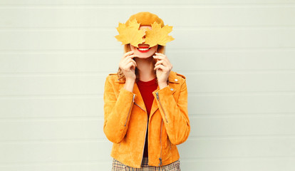 Autumn mood! happy smiling woman holding in her hands yellow maple leaves hiding her eyes over gray wall background