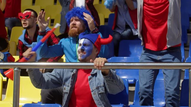 Medium shot of devoted young Caucasian male fans, wearing team colors, wigs and face paint, sitting on tribunes at stadium, agitatedly cheering, then jumping up and down and hugging to celebrate goal