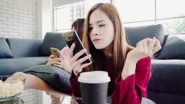 Asian women using smartphone and eating popcorn in living room at home, group of roommate friend enjoy funny moment while lying on the sofa. Lifestyle women relax at home concept.
