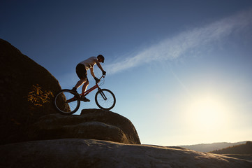 Fototapeta na wymiar Silhouette of man cyclist balancing on trial bicycle, making acrobatic trick on top of mountain on summer sunny evening, blue sky and sunset on background. Concept of extreme sport active lifestyle