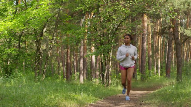 Full tracking shot of slim young Afro-American girl with braided hair, wearing white hoodie and pink shorts, running vigorously along footpath in summer forest on sunny morning