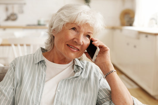 People, modern electronic gadgets, technology and communication. Aged senior woman with short gray hair enjoying nice phone conversation, sitting on sofa, holding mobile at her ear and smiling