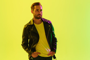 Handsome young stylish man posing isolated over light green background wall with led neon lights dressed in leather jacket.