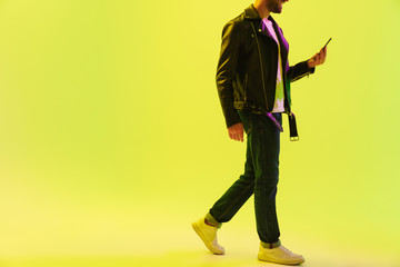 Fototapeta premium Young stylish man isolated over light green background wall with led neon lights dressed in leather jacket using mobile phone.