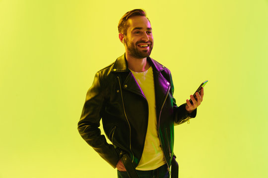 Happy Cheerful Handsome Young Stylish Man Isolated Over Light Green Background Wall With Led Neon Lights Dressed In Leather Jacket Using Mobile Phone.