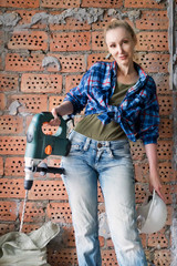 young woman in work clothes and a protective helmet stands next to a brick wall and holds a heavy hammer drill