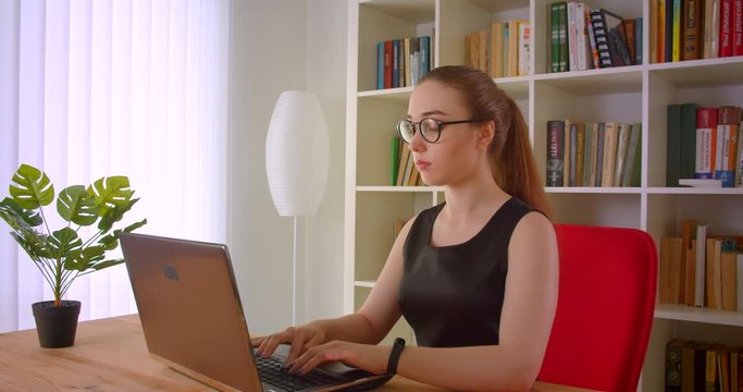 Closeup portrait of young pretty redhead businesswoman in glasses using laptop in office indoors