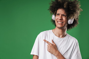 Confused emotional curly guy posing isolated over green wall background listening music with...