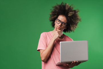 Thinking thoughtful curly guy posing isolated over green wall background using laptop computer.