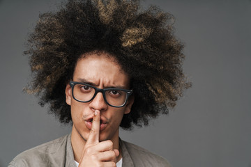 Young curly serious teenage guy posing isolated over grey wall background showing silence gesture.