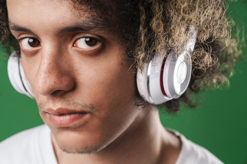 Serious young curly guy posing isolated over green wall background listening music with headphones.