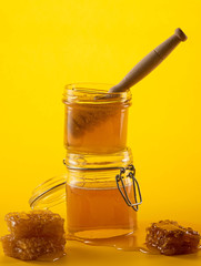 Two jars of honey with honeycombs and a stick on a yellow background