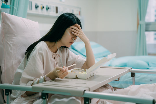 Terminally Ill Asian Woman Wear Pajamas Sitting In Hospital Bed In Lunch Time. Japanese Female Patient Using Chopsticks Eating Healthy Meal In Room. Depressed Young Girl With Hands In Forehead Think