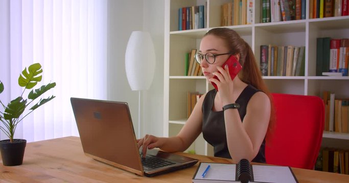 Closeup portrait of young pretty redhead businesswoman in glasses using laptop and having phone call in office indoors