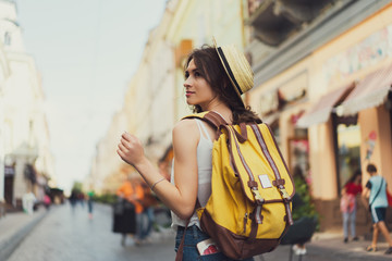 Back view of a young woman traveler with a backpack on her shoulder out sightseeing in a foreign city, stylish female foreigner examines architectural monument during her long-awaited summer vacation - obrazy, fototapety, plakaty