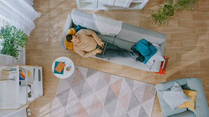 Young Man in Brown Jumper and Grey Jeans Lying Down on a Sofa, Using a Laptop. He is Happy and...