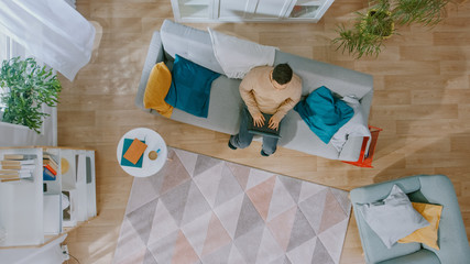 Young Man in Brown Jumper and Grey Jeans Comes and Sits Down on a Sofa, Using a Laptop. Cozy Living Room with Modern Interior with Plants, Table and Wooden Floor. Top View.