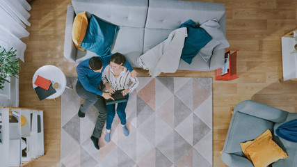 Young Couple is Sitting on a Floor, Emotionally Talking and Using a Laptop. Cozy Living Room with Modern Interior with Carpet, Sofa, Chair, Table, Book Shelf, Plant and Wooden Floor. Top View.