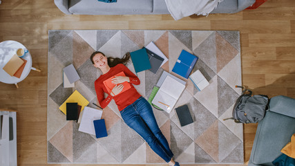Young Girl in Red Coat and Blue Jeans is Lying Down on a Floor, Reading a Notebook. Looks Above, Smiles and Laughs. Cozy Living Room with Modern Interior with Carpet, Workbooks and Backpack. Top View.