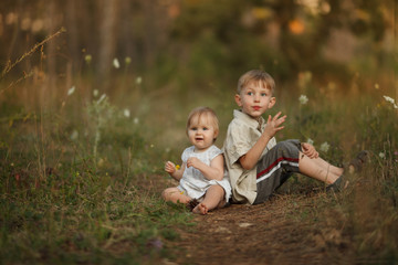 Two cute happy little kids brother and sister sitting with their backs to each other outdoors in the Park. Smiling and laughing looking at camera. The concept of a happy family on holiday in nature.