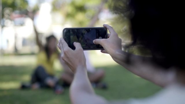 Woman photographing friends with cell phone. Cropped shot of woman holding smartphone and taking pictures of happy young friends in park, selective focus. Technology concept