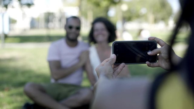 Woman photographing friends with smartphone. Cropped shot of young woman holding cell phone and taking pictures of happy friends in park, selective focus. Technology concept