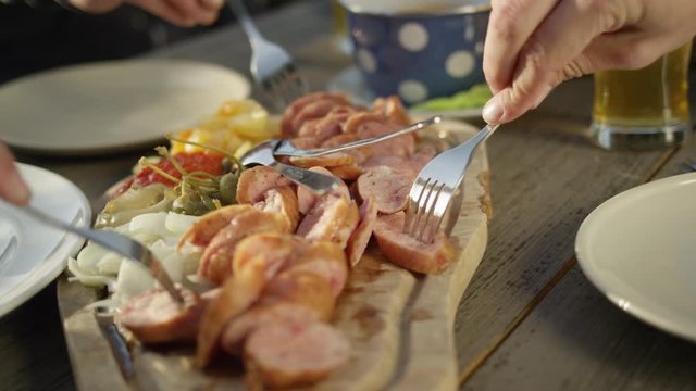 Slow motion, close up friends sit at the picnic table and eat sausage and vegetable on the wooden cutting board