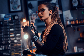 Pretty expirienced tattoo artist in glasses is tuning her new tattoo machine before tattooing session. - obrazy, fototapety, plakaty