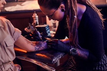 Young focused tattoo master is makining tattoo on client's hand at dark tattoo salon.