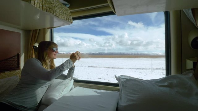 CLOSE UP: Cheerful Caucasian woman taking photos of the snowy Tibetan landscape during a long train ride. Young female traveler having fun observing the Himalaya while staying in her first class cabin