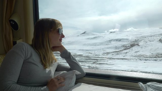 SLOW MOTION CLOSE UP: Cheerful young woman observes the scenic snowy Himalaya during a train ride. Female traveler daydreams while crossing the snow covered Tibetan plains in first class sleeper train