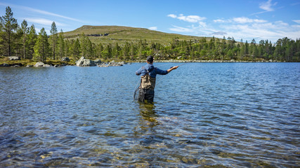 Fototapeta na wymiar Angler mit Angel in Wathose im Wasser beim Fliegenfischen bei Sonne vor Wald und Berg bei Sonne