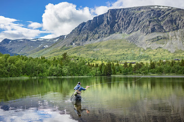 Angler beim Fliegenfischen mit Angelroute im Wasser stehend mit Wathose im See vor Bäumen und Berg