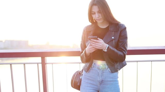 Portrait of a beautiful young woman against the backdrop of the city sunset. ide view of young hipster girl wearing leather jacket and chatting with friends at social networks via smartphone