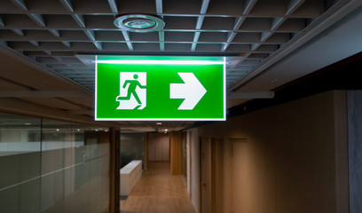 Green fire escape sign hang on the ceiling in the office. - obrazy, fototapety, plakaty