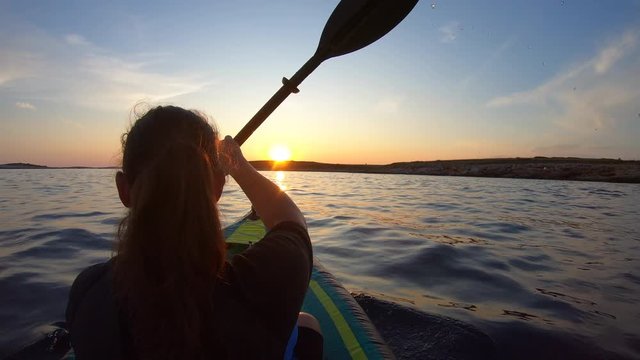 Right behind shot of a woman paddling her kayak at sea into the setting sun