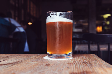 A glass of beer with foam on a wooden table. A sip of refreshing brown drink in the bar. Loneliness.