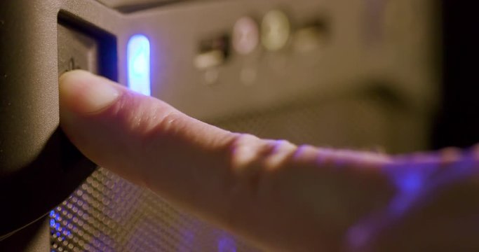 A finger of a man pushes the power button of a computer which illuminates a blue light indicating it is booting up. Macro Shot