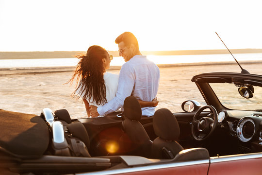 Photo from back of happy multiethnic couple hugging together while standing by car on beach