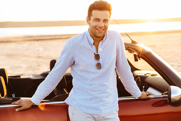 Photo of brunette caucasian man standing by convertible car on beach at sunrise