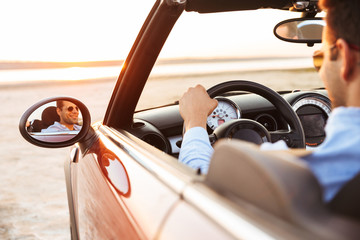 Photo of joyful handsome man driving convertible car by seaside at sunrise